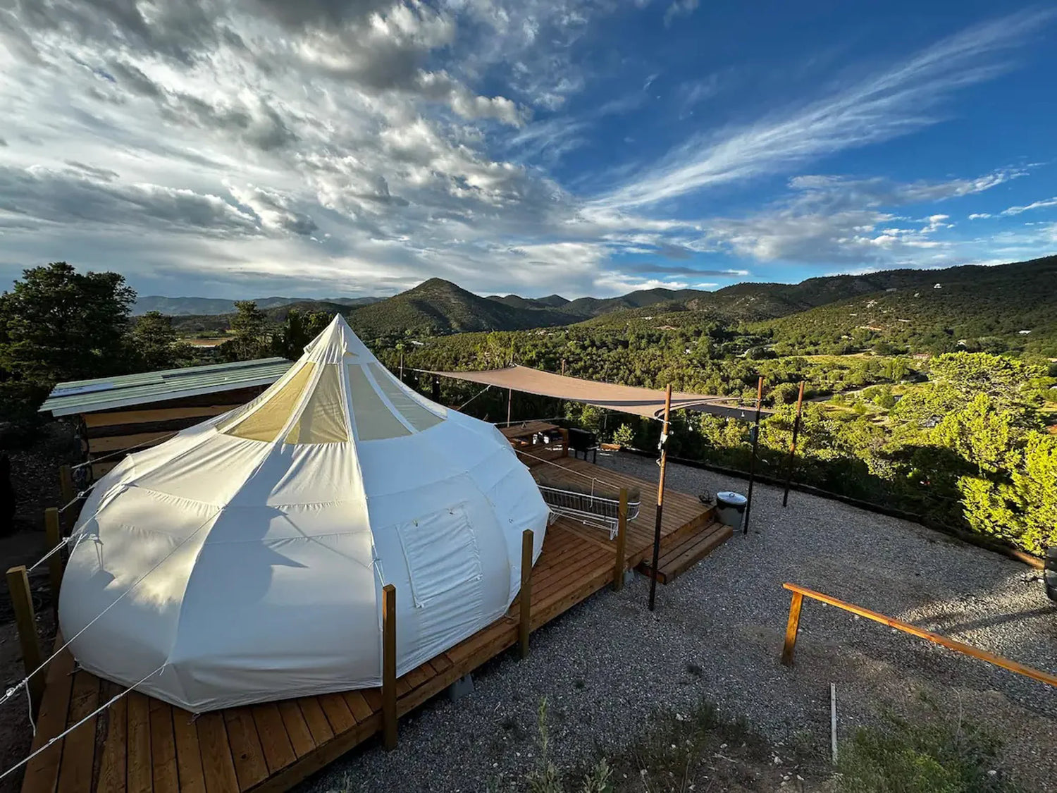 White tent-like structure with wooden deck and surrounding greenery under a blue sky.
