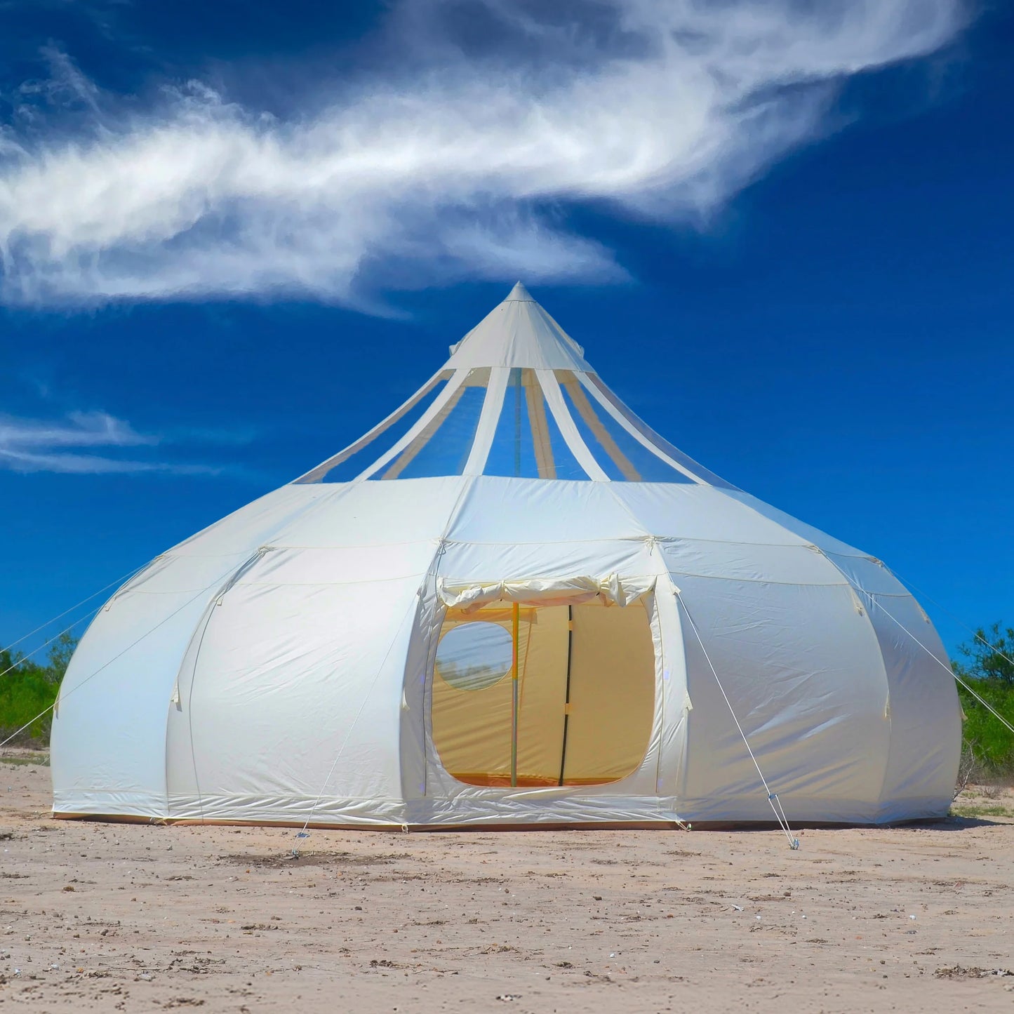 Large white tent with a yellow door on a sandy ground under a blue sky with clouds.