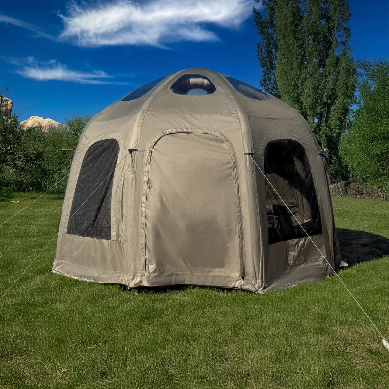 Beige dome tent on grass with trees and blue sky in the background