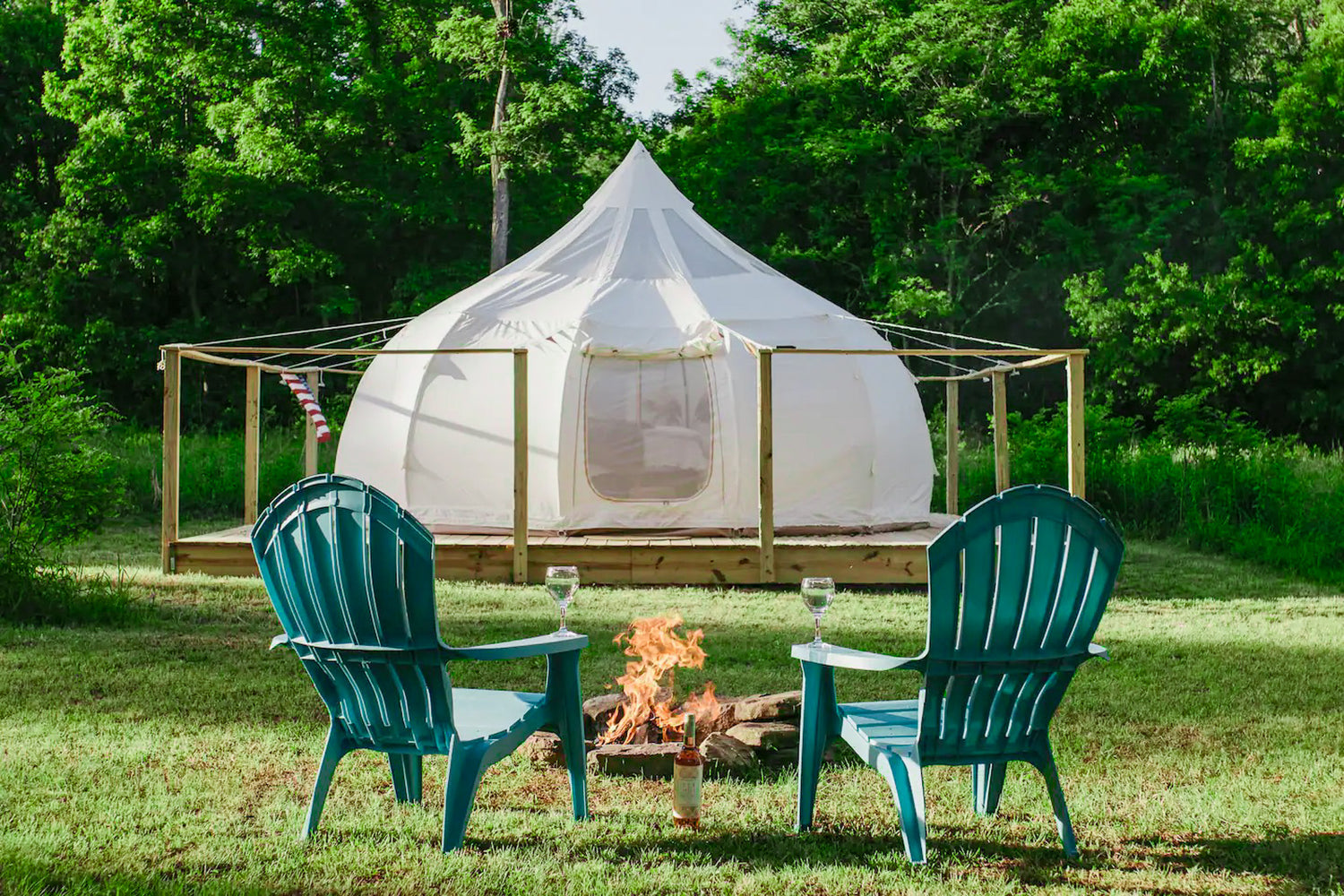 White tent with green Adirondack chairs and a fire pit in a forest setting