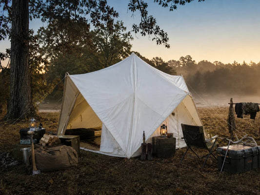A canvas glamping tent set up as a hunting basecamp at dawn with a wood stove pipe and gear nearby