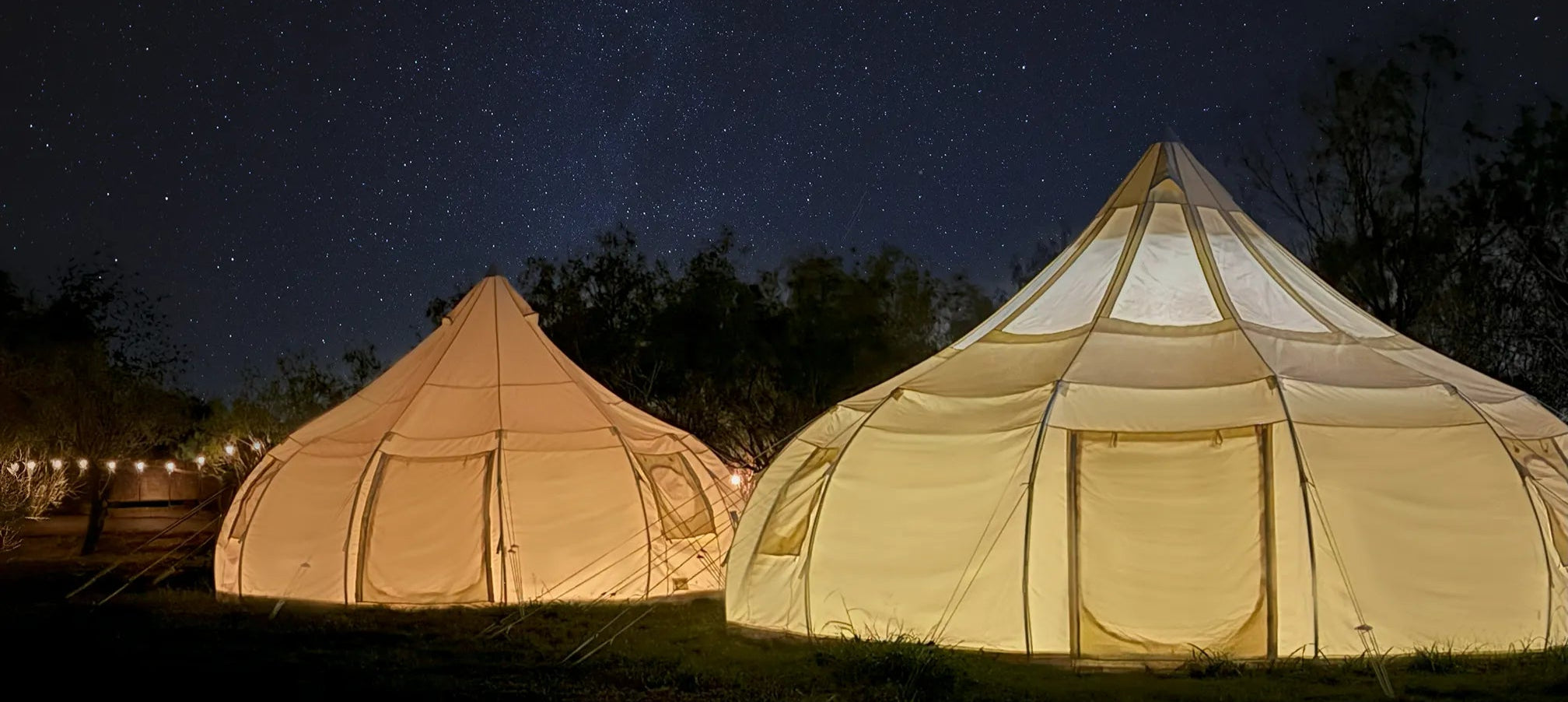 Two illuminated tents under a starry night sky with trees in the background.