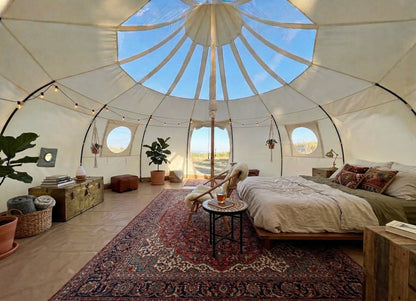 Furnished Astral glamping tent interior with queen bed, oriental rugs, string lights, plants, wooden furniture, and natural light through skylight ceiling